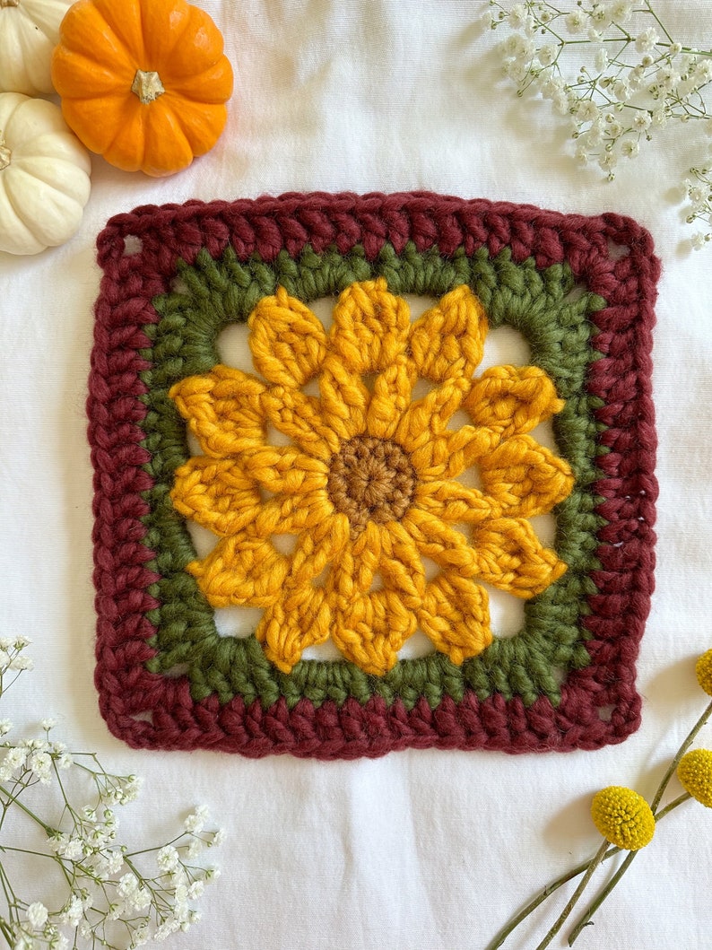 May include: A crocheted sunflower square with a burgundy border, a green inner border, and a yellow sunflower in the center.