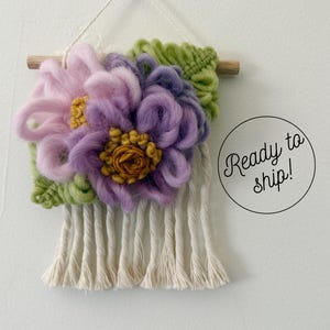 May include: A wall hanging featuring soft, looped yarn flowers in shades of light pink and purple with yellow centers, accented by green macrame leaves. The piece is suspended from a wooden dowel with white cord fringe and includes the text Ready to ship!