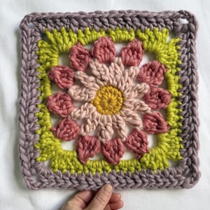 May include: A crocheted square with a flower design in shades of pink, yellow, and green. The flower has a center of yellow yarn and petals of pink and light pink yarn. The square is bordered with a dark purple yarn.