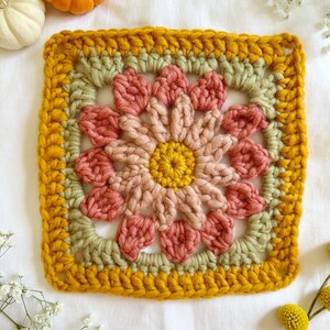 May include: A crocheted square with a flower design in shades of pink, green, and yellow. The square is surrounded by a yellow border.