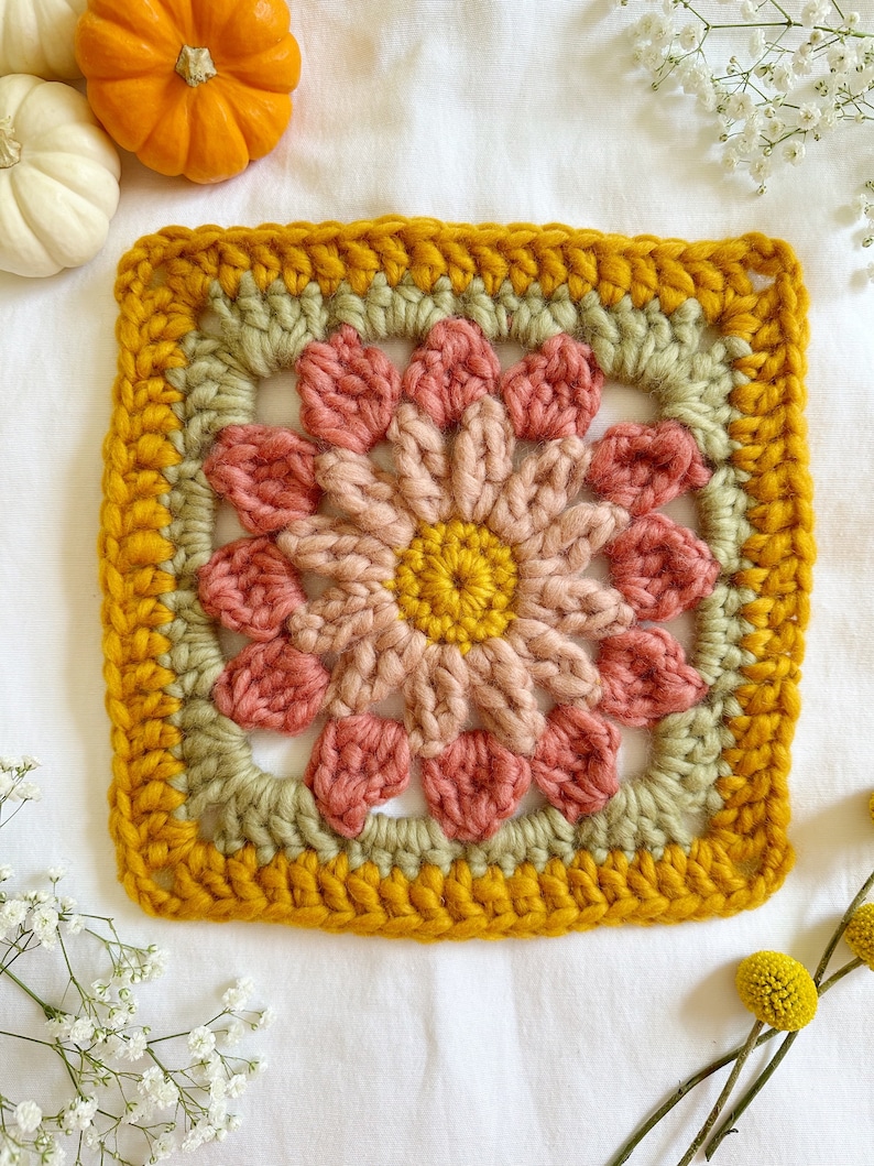 May include: A crocheted square with a flower design in shades of pink, green, and yellow. The square is surrounded by a yellow border.