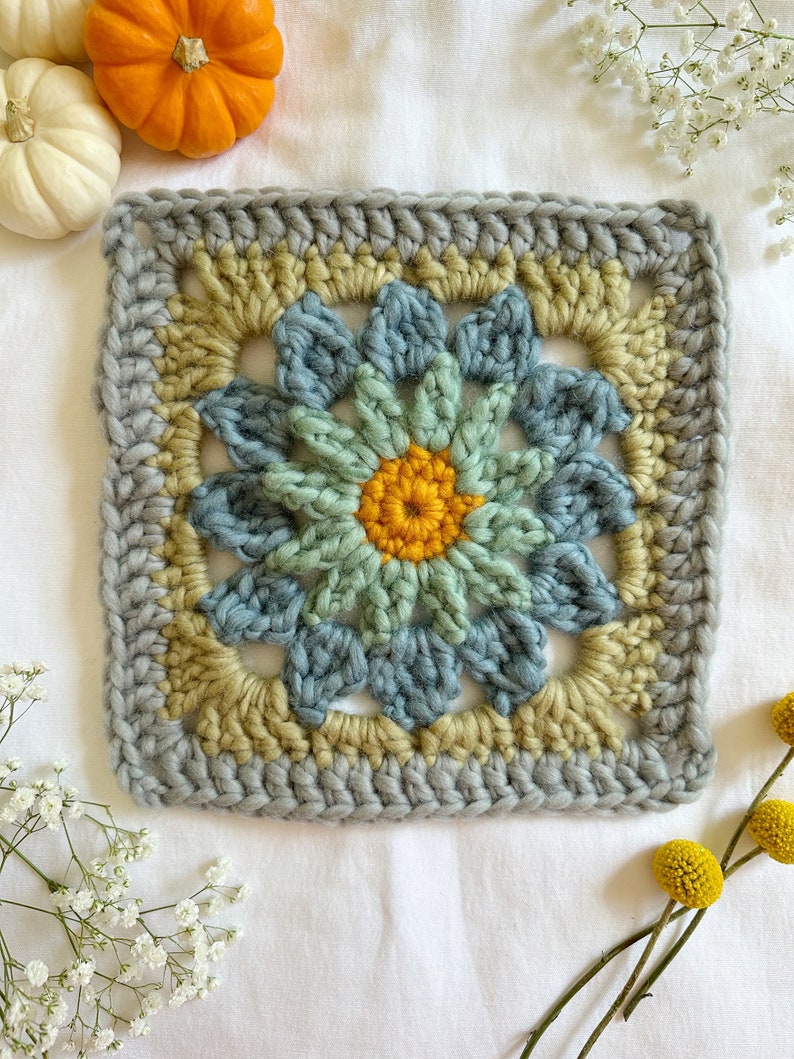 May include: A crocheted square with a flower design in shades of blue, green, and yellow. The flower has a center of yellow yarn and petals in shades of blue and green.