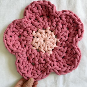 May include: A handmade, pink flower-shaped trivet with a lighter pink center. The trivet is made of thick, braided fabric, and is designed to protect surfaces from hot items. The flower has five petals.