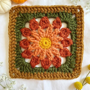 May include: A crocheted square with a flower design in shades of orange, red, and yellow. The flower is surrounded by a green border and a brown outer border.