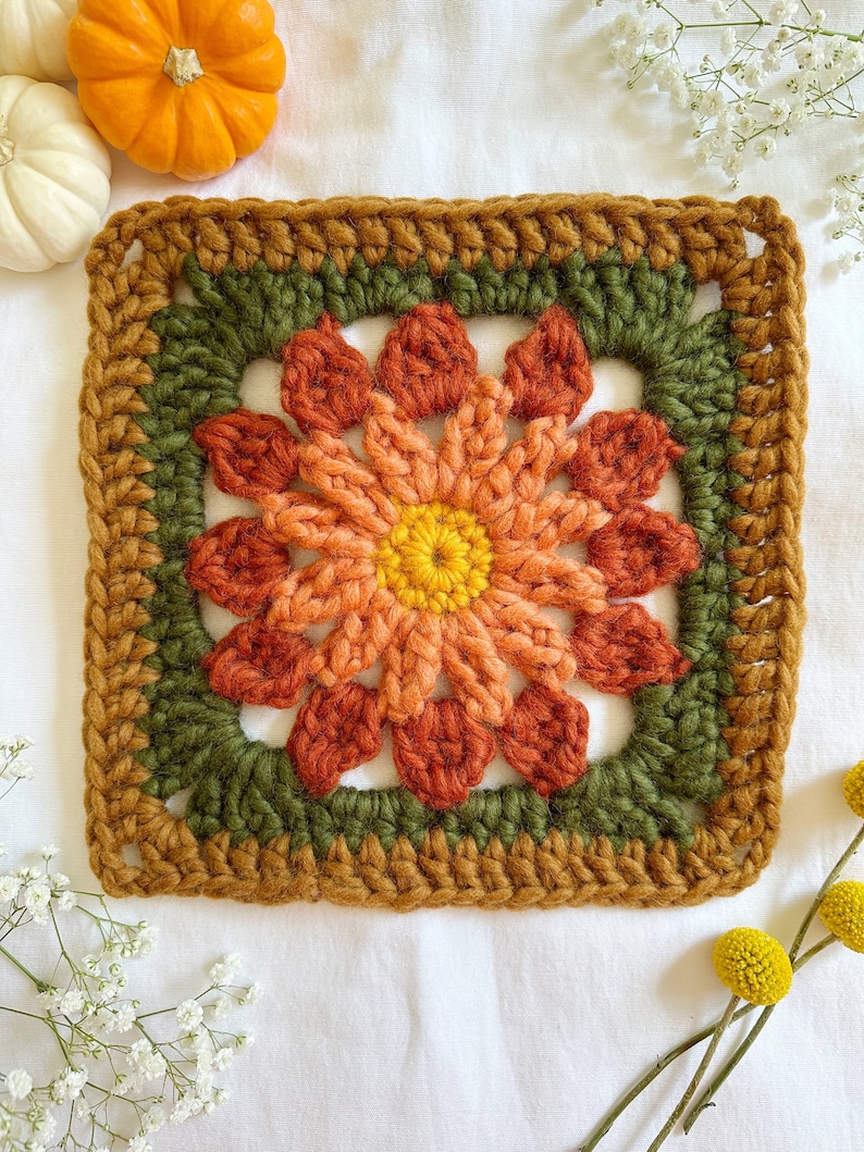 May include: A crocheted square with a flower design in shades of orange, red, and yellow. The flower is surrounded by a green border and a brown outer border.
