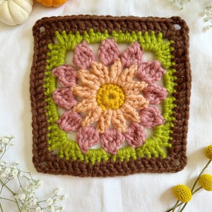 May include: A crocheted square with a flower design in shades of brown, green, pink, and yellow. The square is surrounded by a brown border.