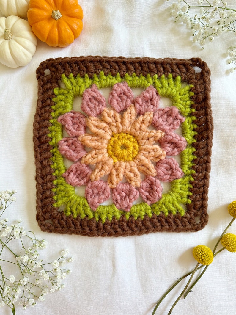 May include: A crocheted square with a flower design in shades of brown, green, pink, and yellow. The square is surrounded by a brown border.