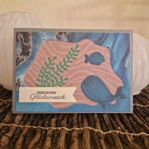 Könnte beinhalten: Eine handgemachte Grußkarte mit einem blauen Wal, einem kleinen Fisch und einer grünen Pflanze auf einem rosa-blauen Hintergrund. Die Karte hat ein weißes Banner mit dem Text "Herzlichen Glückwunsch".