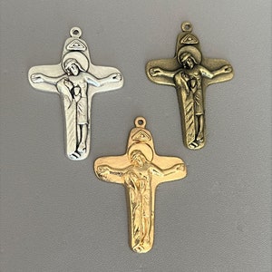 May include: Three metal crucifix pendants in silver, bronze, and gold. Each cross features a detailed depiction of Jesus Christ. The pendants have a loop at the top for attaching to a chain or cord. The background is a solid gray.
