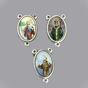 May include: Three oval religious pendants with silver-colored frames. Each pendant features a different saintly image in vibrant colors. The pendants have small loops for stringing or attaching to other items. The background is a solid gray.