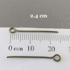 Op de afbeelding: Twee goudkleurige kopspelden, elk 2,4 cm lang, met een lus aan het ene uiteinde. De spelden liggen op een wit oppervlak met een liniaal die in millimeters is gemarkeerd.