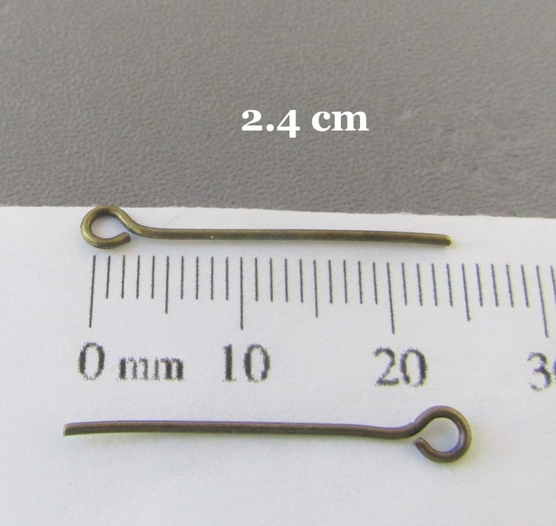 Op de afbeelding: Twee goudkleurige kopspelden, elk 2,4 cm lang, met een lus aan het ene uiteinde. De spelden liggen op een wit oppervlak met een liniaal die in millimeters is gemarkeerd.