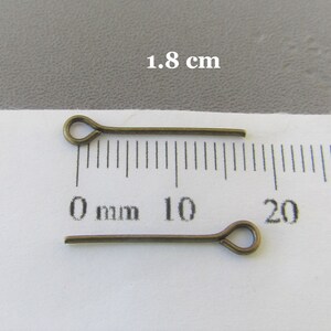 Op de afbeelding: Twee 1,8 cm lange bronzen kopspelden met een lus aan het ene uiteinde. De spelden liggen op een wit oppervlak met een liniaal die in millimeters is gemarkeerd.