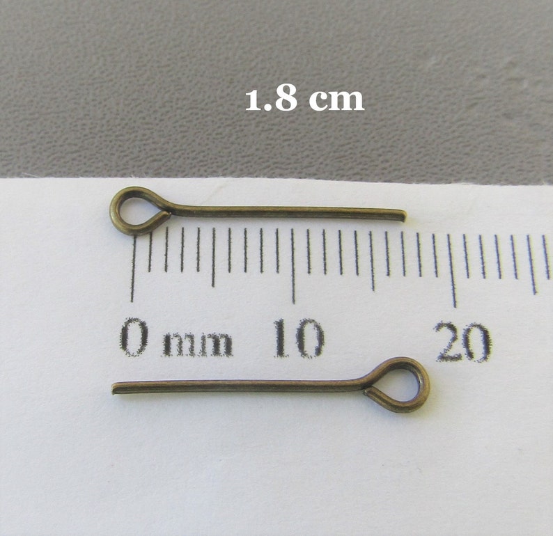 Op de afbeelding: Twee 1,8 cm lange bronzen kopspelden met een lus aan het ene uiteinde. De spelden liggen op een wit oppervlak met een liniaal die in millimeters is gemarkeerd.