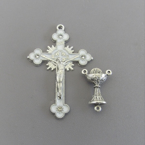 May include: A silver-toned crucifix with a white enamel finish and rhinestone accents, alongside a matching chalice. The cross features a detailed depiction of Jesus Christ. The chalice has a detailed grape design.
