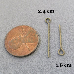 Op de afbeelding: Twee goudkleurige kopspelden, elk met een lus aan de bovenkant. De spelden zijn respectievelijk 2,4 cm en 1,8 cm lang. De spelden worden naast een Amerikaanse penny getoond ter vergelijking van de grootte.