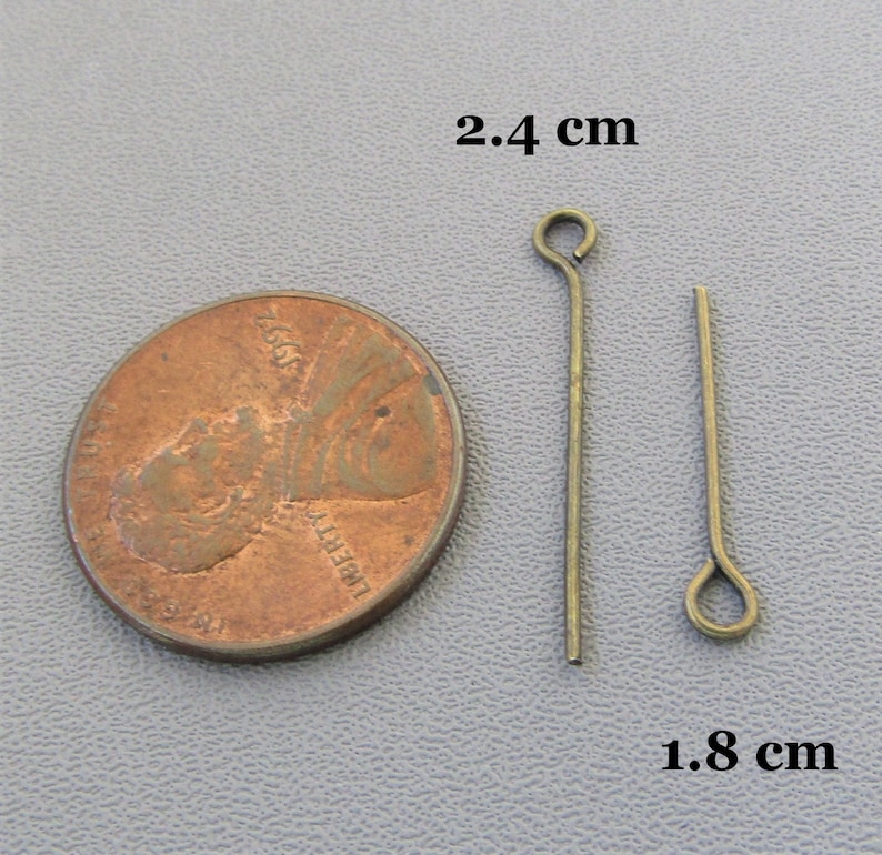 Op de afbeelding: Twee goudkleurige kopspelden, elk met een lus aan de bovenkant. De spelden zijn respectievelijk 2,4 cm en 1,8 cm lang. De spelden worden naast een Amerikaanse penny getoond ter vergelijking van de grootte.