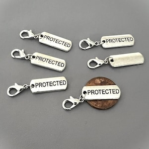 May include: Silver-toned metal charms with the word "PROTECTED" engraved on rectangular tags. Each charm has a lobster clasp for easy attachment. One charm is placed on a penny. The charms are displayed on a gray surface.