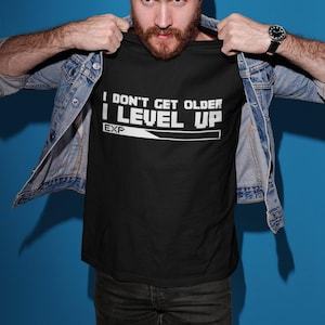 May include: Black t-shirt with the text "I DON'T GET OLDER I LEVEL UP" in white. The shirt is worn with a denim jacket, a grey cap, and dark jeans. A watch is visible on the wrist.