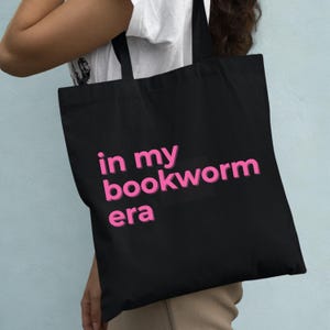 Op de afbeelding: Zwarte draagtas met de roze tekst "in my bookworm era". De tas heeft twee zwarte handvatten en wordt door een persoon vastgehouden. De achtergrond is lichtblauw.