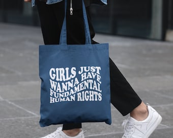 May include: A blue tote bag with the text "Girls Just Wanna Have Fundamental Human Rights" in white. The bag is being held by a person wearing a denim shirt and black trousers.