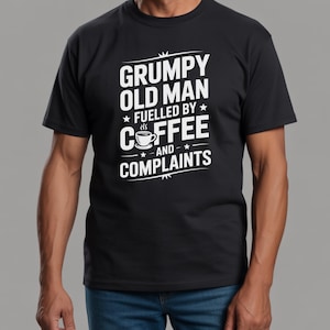 Op de afbeelding: Zwart t-shirt met witte tekst: "GRUMPY OLD MAN FUELLED BY COFFEE AND COMPLAINTS". Het shirt heeft een koffiekopje en sterren. Het shirt wordt door een persoon gedragen.