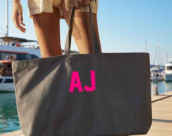 May include: A large gray tote bag with the hot pink letters "AJ" printed on the front. The bag has two long handles and is being held by a person on a wooden dock near the water.