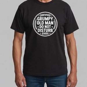 Könnte beinhalten: Schwarzes T-Shirt mit Rundhalsausschnitt und weißem, kreisförmigem Aufdruck. Der Aufdruck lautet "CERTIFIED GRUMPY OLD MAN DO NOT DISTURB (EVER)" in einer kräftigen Schrift, umgeben von einem Lorbeerkranz. Das T-Shirt ist aus weichem Material.