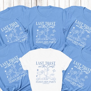 May include: A collection of short-sleeved t-shirts in light blue and white. Each shirt features a white graphic print with the text "LAST TOAST on the Coast" above illustrations of three cocktails, seashells, and stars, with "FLISS'S HEN PARTY CYPRUS 2027" below.
