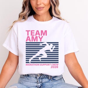 Könnte beinhalten: Weißes T-Shirt mit dem pinkfarbenen Text "TEAM AMY" über einem blau-weiß gestreiften Design mit einer Läuferfigur. Darunter steht der Text "MARATHON SUPPORT CREW 2026".