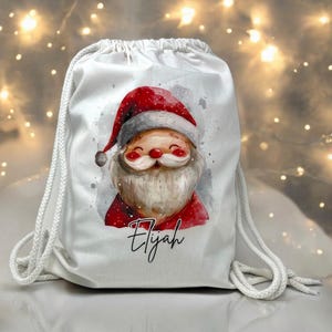 Könnte beinhalten: Weißer Kordelzugbeutel mit einer Aquarellillustration des Weihnachtsmanns mit rotem Hut und Mantel. Die Tasche hat einen weißen Kordelzugverschluss und der Name "Elijah" ist in Kursivschrift unter der Weihnachtsmann-Illustration geschrieben. Hintergrund mit Bokeh-Lichtern.