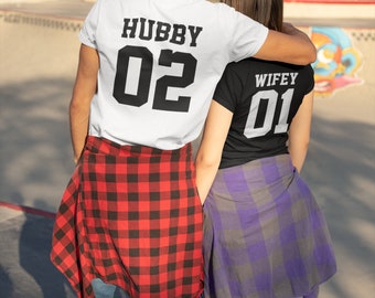 Może przedstawiać: Para nosząca dopasowane koszulki z napisem "Hubby 02" i "Wifey 01" na plecach. Mężczyzna ma na sobie białą koszulkę z czarnym napisem, a kobieta czarną koszulkę z białym napisem. Oboje mają na sobie kratowane koszule na swoich koszulkach.