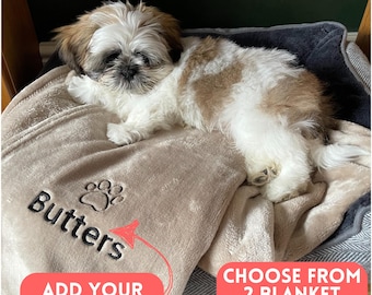 May include: A small, fluffy dog rests on a beige blanket embroidered with the name "Butters" and a paw print. The blanket is part of a pet bed set. Text on the image reads "Add your dog's name" and "Choose from 2 blanket colors."