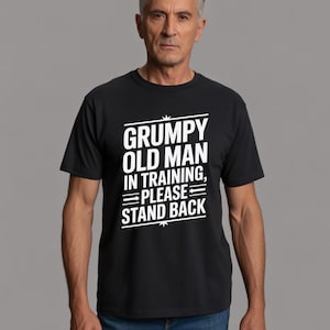 以下が含まれることがあります： 「GRUMPY OLD MAN IN TRAINING, PLEASE STAND BACK」の白文字がプリントされた黒いTシャツ。Tシャツを着ている人は白髪で、表情はニュートラルです。背景は無地のグレーです。