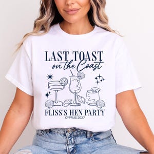 May include: A white short-sleeve t-shirt featuring a dark blue graphic print. The design includes three cocktail glasses, seashells, and star icons. The text reads: LAST TOAST on the Coast, FLISS'S HEN PARTY, and CYPRUS 2027 in a mix of fonts.