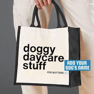 May include: A white tote bag with black trim and handles. The bag features the words "doggy daycare stuff" in large black letters. Below the text is the name "For Butters". A blue box says "Add your dog's name".
