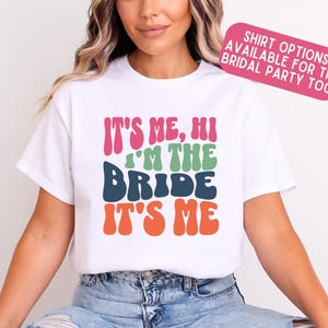 Op de afbeelding: Wit T-shirt met de tekst "It's Me, Hi I'm the Bride It's Me" in kleurrijk retro-lettertype.