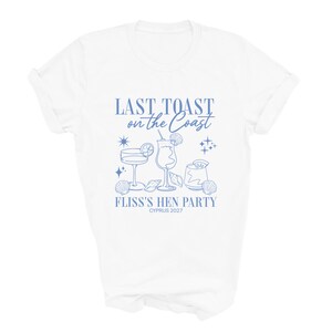 May include: A white short-sleeve t-shirt featuring a blue graphic print with the text "LAST TOAST on the Coast", "FLISS'S HEN PARTY", and "CYPRUS 2027". The design includes illustrations of three cocktail glasses, seashells, and stars.