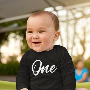 May include: A black long-sleeve shirt featuring the word One printed in a white cursive script font across the chest. The garment has a round neckline and long sleeves, providing a simple and clean design for a first birthday celebration outfit.