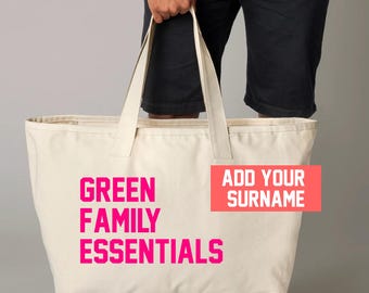 May include: Cream-colored canvas tote bag with the text "GREEN FAMILY ESSENTIALS" in bright pink. A rectangular pink box with the text "ADD YOUR SURNAME" is on the right side of the bag. The bag has long handles.