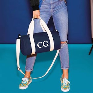 Peut inclure: Un sac de sport bleu marine avec des sangles blanches et les initiales "CG" brodées sur le devant. Le sac est porté par une personne vêtue d'un jean bleu.