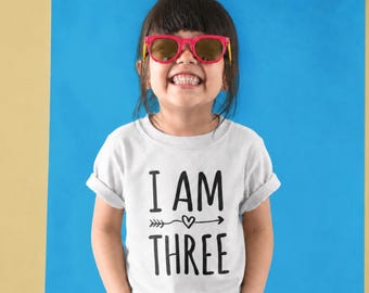 Könnte beinhalten: Ein weißes kurzärmeliges T-Shirt mit dem schwarzen Text I AM THREE und einem kleinen Herz- und Pfeilmotiv zwischen den Wörtern. Das T-Shirt wird mit einer leuchtend pinken Sonnenbrille getragen und das Design ist in einer serifenlosen Schrift.
