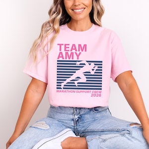 Op de afbeelding: Lichtroze t-shirt met de tekst "TEAM AMY" in roze boven een witte hardloper. Daaronder staat de tekst "MARATHON SUPPORT CREW 2026" in het wit. Het t-shirt wordt door een persoon gedragen.