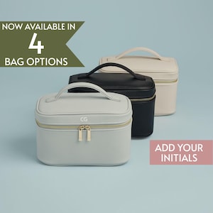 May include: Three cosmetic bags in light gray, black, and cream colors, each with a top handle and gold-colored zipper. The light gray bag has the initials "CG" on the front. Text reads "NOW AVAILABLE IN 4 BAG OPTIONS" and "ADD YOUR INITIALS".
