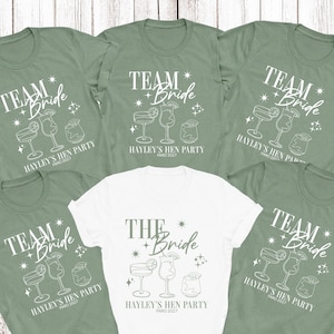May include: Five short-sleeved crew neck t-shirts, four in sage green and one in white. Each shirt features a white graphic print with the text "TEAM BRIDE" or "THE BRIDE", "HAYLEY'S HEN PARTY", and "FARO 2027" alongside illustrations of cocktail glasses.