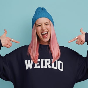 May include: A woman with pink hair and a blue beanie is wearing a navy blue sweatshirt with the word "WEIRDO" printed in white on the front. She is pointing at herself with both hands.