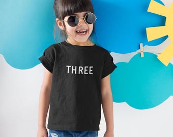 Könnte beinhalten: Ein schwarzes Kurzarm-T-Shirt mit dem Wort THREE in weißen Großbuchstaben auf der Brust. Das Oberteil hat einen Rundhalsausschnitt und umgekrempelte Ärmel, kombiniert mit einer blauen Jeans und einer runden schwarzen Sonnenbrille.