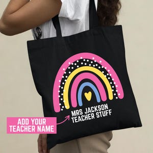 May include: Black tote bag featuring a colorful rainbow design with the text "MRS JACKSON TEACHER STUFF". The rainbow includes pink, yellow, and blue arches with white polka dots. A pink rectangle with the text "ADD YOUR TEACHER NAME" is also visible.