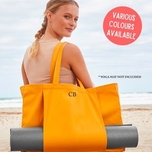 Op de afbeelding: Een gele canvas draagtas met de initialen "CB" geborduurd op de voorkant. De tas is groot genoeg om een yogamat in te bewaren, die gedeeltelijk zichtbaar is op de afbeelding. De tekst "Various Colours Available" en "Yoga Mat Not Included" is ook zichtbaar op de afbeelding.