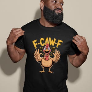 Puede incluir: Camiseta negra con un dibujo de un pollo de dibujos animados y el texto "F-CAW-F" en amarillo. El pollo tiene una cresta roja y un pico amarillo, con las alas extendidas. La camiseta la sostiene una persona.